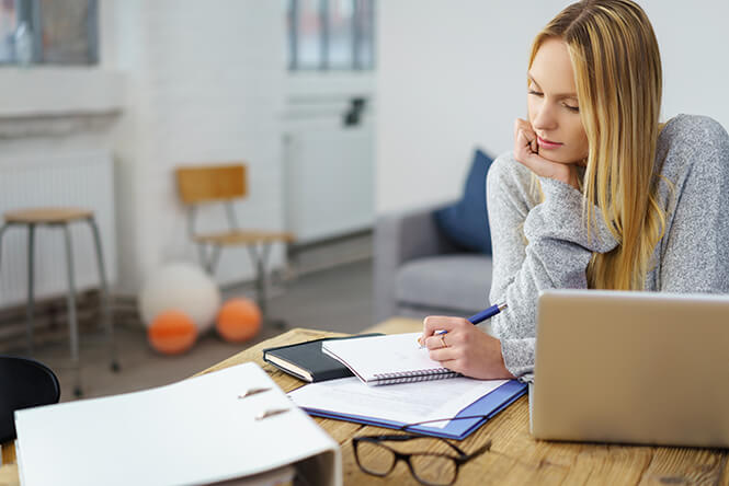 Junge Frau sitzt an einem Schreibtisch mit Laptop, Ordner und Notizbuch und schreibt konzentriert – Lernumgebung im Homeoffice-Stil.