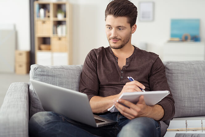 Mann sitzt auf einem Sofa, arbeitet mit Laptop und Notizblock und macht sich konzentriert Notizen – Lernsituation in entspannter Wohnatmosphäre.